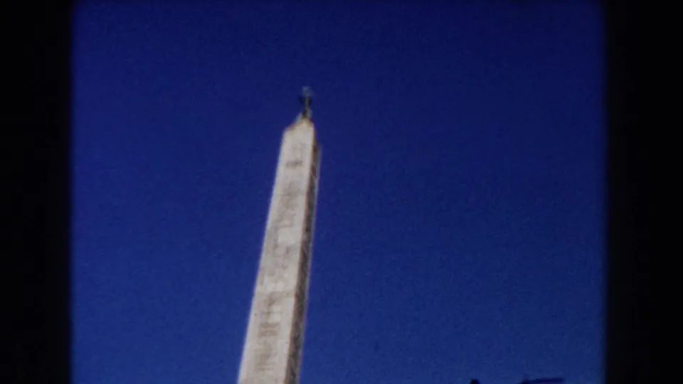 1959 Rome frame showing the upper portion of a stone obelisk with faint inscriptions and a metal finial against blue sky.