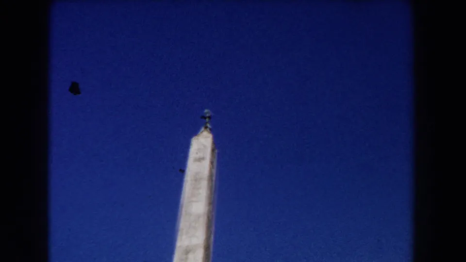 1959 Rome home-movie frame showing a slightly lower, distant view of the obelisk with its finial and darkened 8mm frame edge.