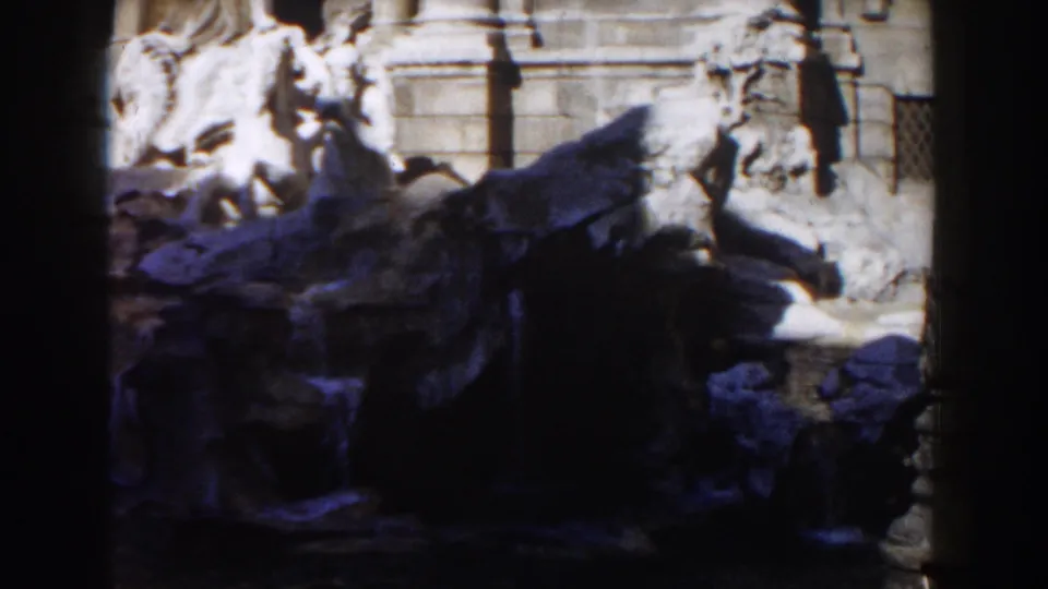 1959 Rome home-movie frame showing a stone sculptural group or fountain with figures near an ornate architectural wall.