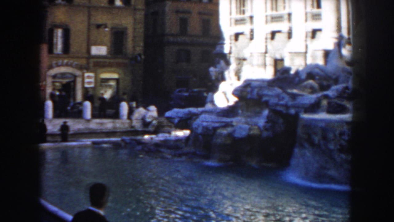 1959 Rome home-movie frame showing a public fountain, sculpted rockwork, storefront facades, and pedestrians near the water.