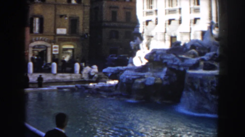 1959 Rome home-movie frame showing a public fountain, sculpted rockwork, storefront facades, and pedestrians near the water.