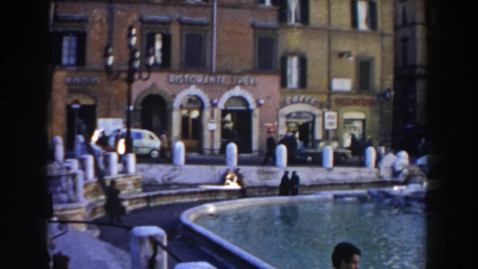 1959 Rome home-movie frame looking across the fountain toward storefronts, a small white car, pedestrians, bollards, and older facades.