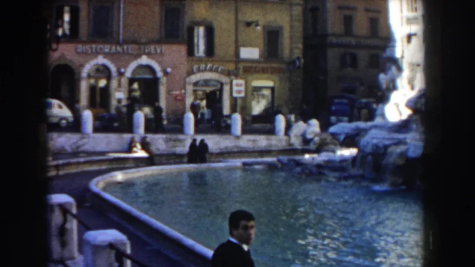 1959 Rome frame showing the fountain basin, restaurant and cafe signage, pedestrians near the water, parked vehicles, and a suited man at the bottom edge.