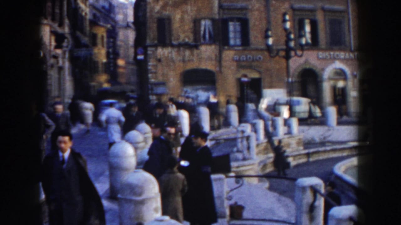 Pedestrians walk beside stone posts near shops and a parked light-colored car.