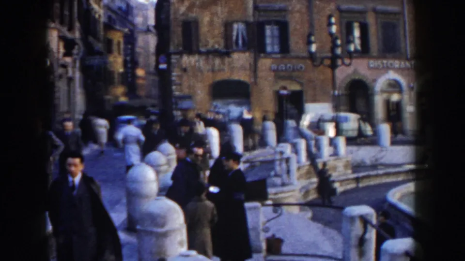 1959 Rome street scene near Trevi Fountain with pedestrians, stone bollards, Italian storefront signs, and a light-colored car.
