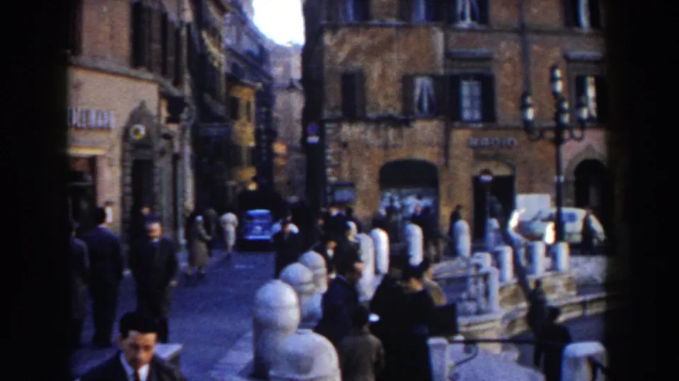 1959 Rome home-movie view of a busy walkway by Trevi Fountain, with pedestrians, railings, bollards, and shop signs.