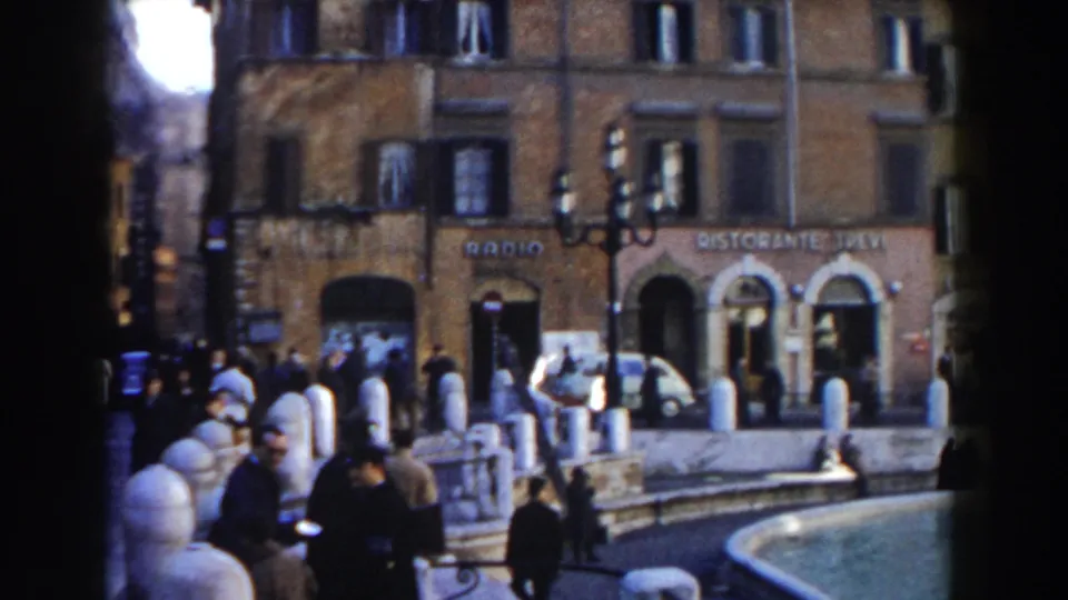 1959 Rome frame showing the Trevi Fountain edge, pedestrians in dark clothing, storefront signage, and a light-colored car nearby.