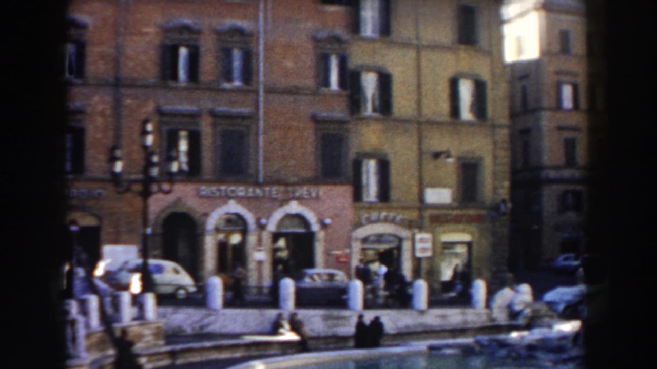 Street-level view of restaurant storefronts, pedestrians, cars, bollards, and building facades.