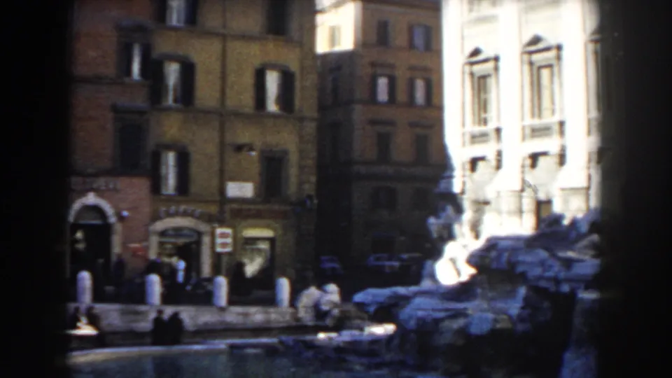1959 Rome footage frame showing a panning view past storefronts, pedestrians, parked cars, fountain stonework, and sunlit architecture.