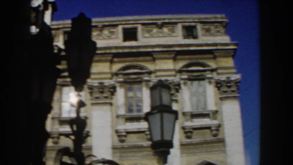 1959 Rome home-movie frame of an ornate historic facade with shuttered windows, carved stone details, a streetlamp, and clear sky.