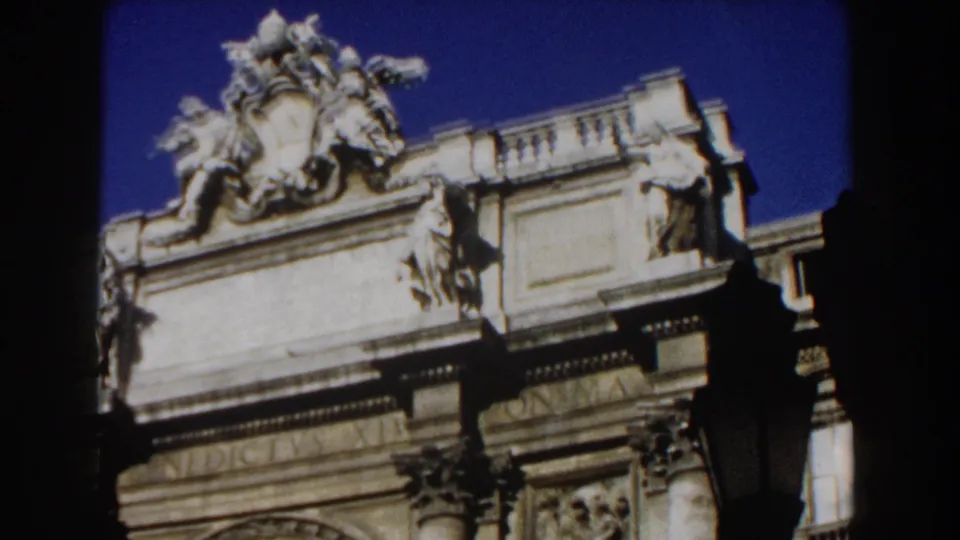 1959 Rome 8mm frame with a sculptural crest, carved figures, balustrade, and upper stone facade seen behind foreground shadows.
