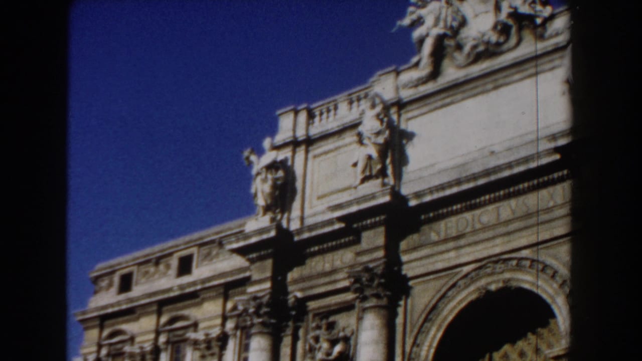 1959 Rome home-movie frame showing a sunlit monumental stone facade with sculptural figures, columns, and an arched opening.