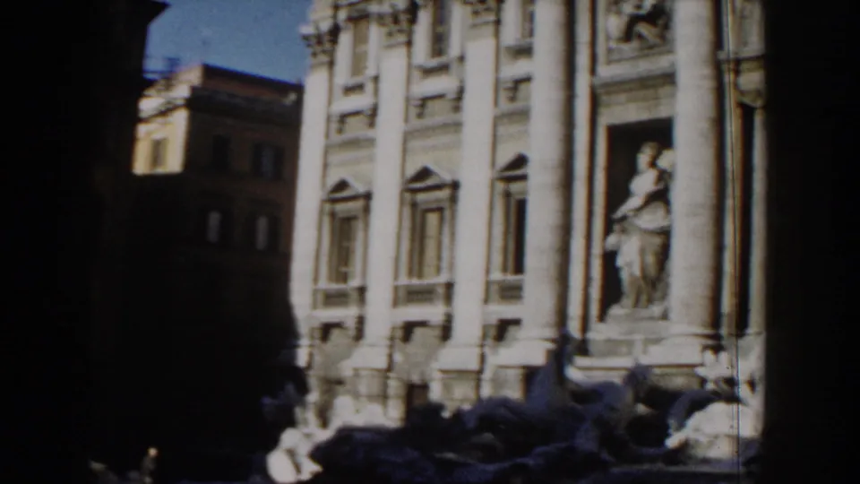 1959 Rome tourist-film frame showing the lower portion of a monumental facade with columns, statue niche, fountain forms, and shadowed foreground.
