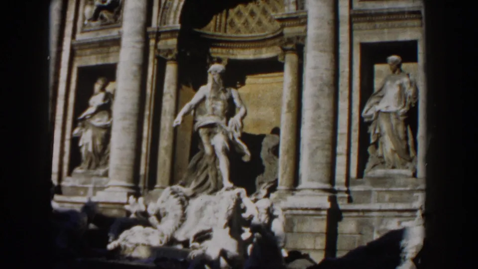 1959 Rome travel footage frame showing Trevi Fountain sculptures, tall columns, carved facade panels, and deep shadow.