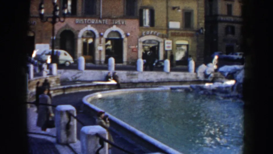 1959 Rome view of the Trevi Fountain area with pedestrians near the basin, white bollards, storefronts, and a small white car.