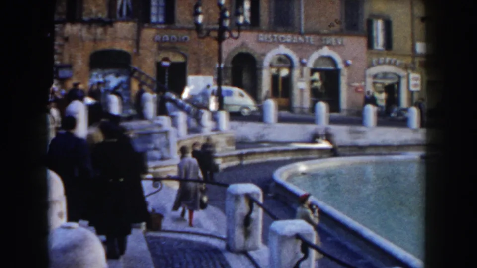 1959 Rome home-movie frame showing people beside the Trevi Fountain, stone posts, street facades, and visible restaurant signage.