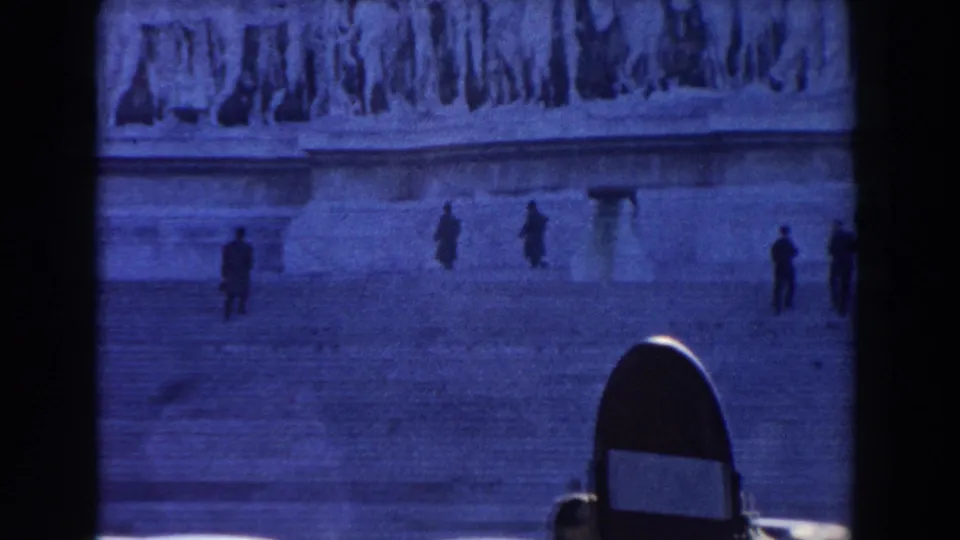 Rome home-movie frame showing a distant view of the Altar of the Homeland area with relief sculpture, plaza, and scattered figures.
