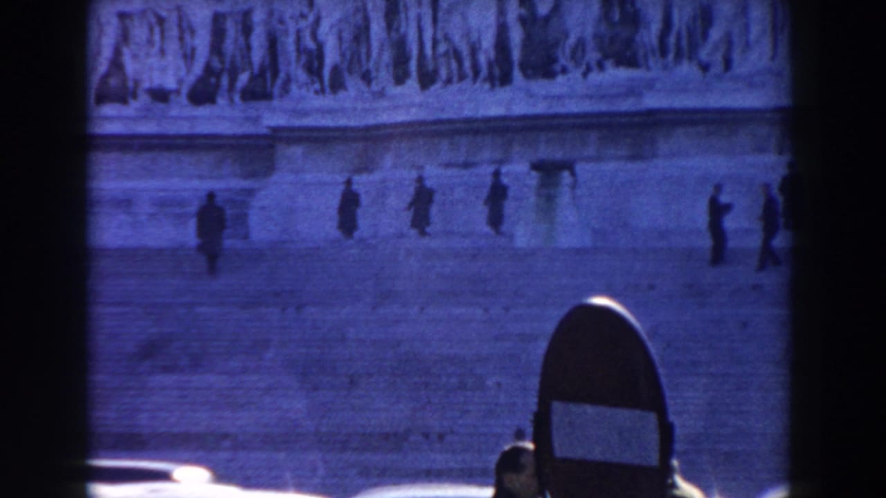 1959 Rome home-movie frame with distant figures on broad stone steps beneath sculpted reliefs, seen through a dark foreground frame.