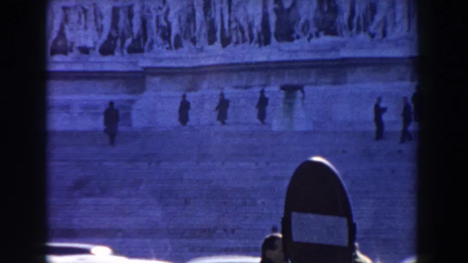1959 Rome home-movie frame with distant figures on broad stone steps beneath sculpted reliefs, seen through a dark foreground frame.