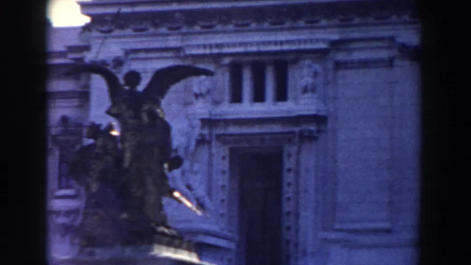 1959 Rome frame with a winged angel sculpture seen against a classical facade with a large doorway and decorative stonework.