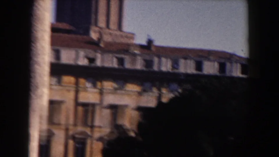 1959 Rome 8mm view of a classical urban facade with shuttered windows, a roofline, palm trunk, and heavy foreground shadow.