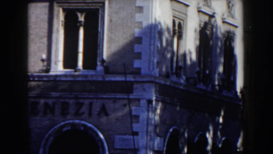 1959 Rome frame of a stone corner building with arched windows, columns, tree shadows, and partial commercial lettering.