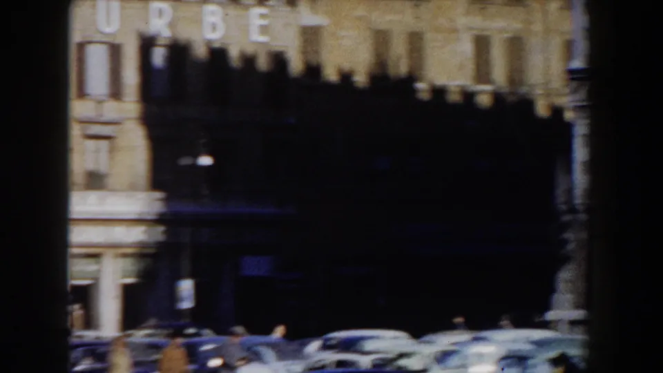 Rome street scene with a prominent building facade, dark awning, older cars, and pedestrians visible at a distance.