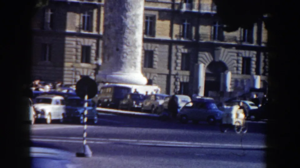 1959 Rome street view with a tall column, rounded period cars, pedestrians, and a masonry building facade.