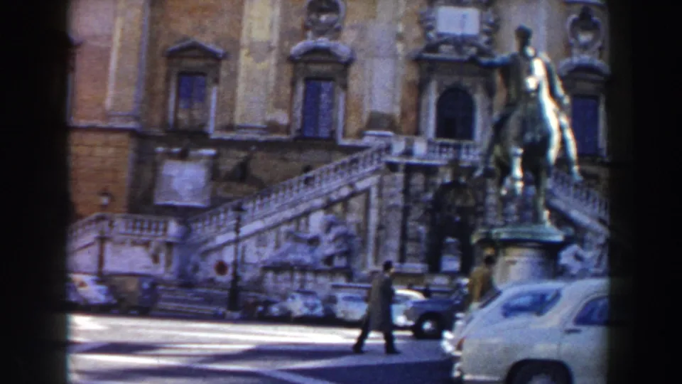 1959 Rome home-movie frame with an equestrian statue near a grand staircase, pedestrians, and rounded cars in sunlight.