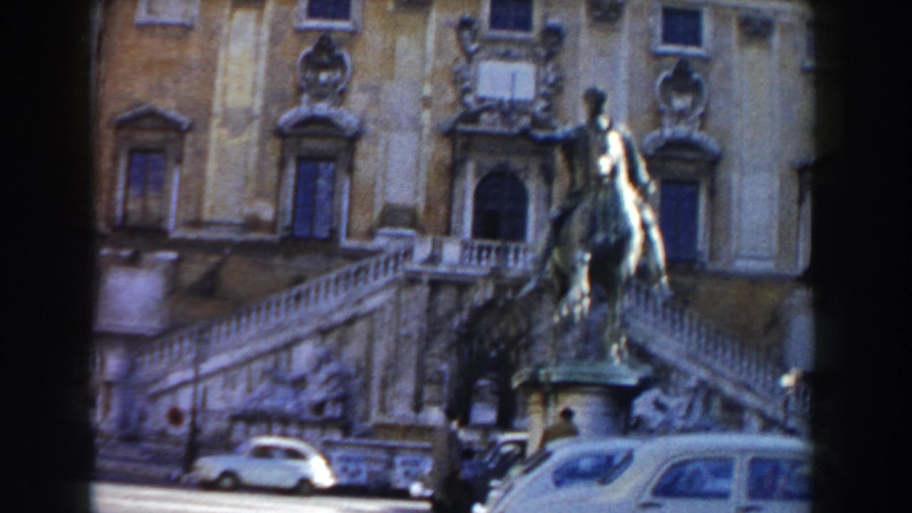 1959 Rome 8mm frame showing an equestrian statue in front of a grand stone facade, with stairs, cars, and a pedestrian in view.