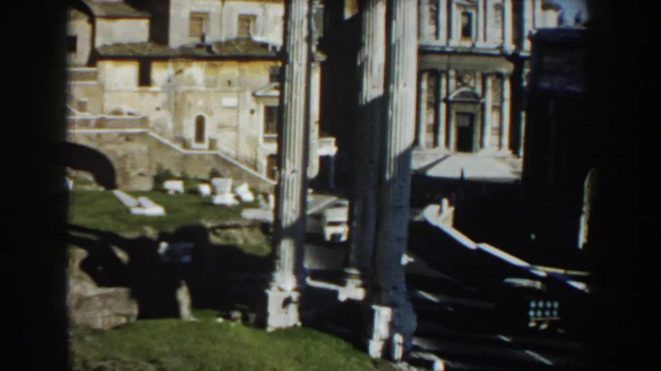 1959 Rome 8mm frame showing sunlit archaeological ruins with columns, stonework, grass, and nearby historic buildings.