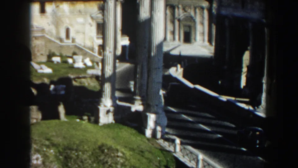 1959 Rome 8mm view across ruins, with tall columns, grassy ground, stone remains, and a nearby building facade in bright sun.