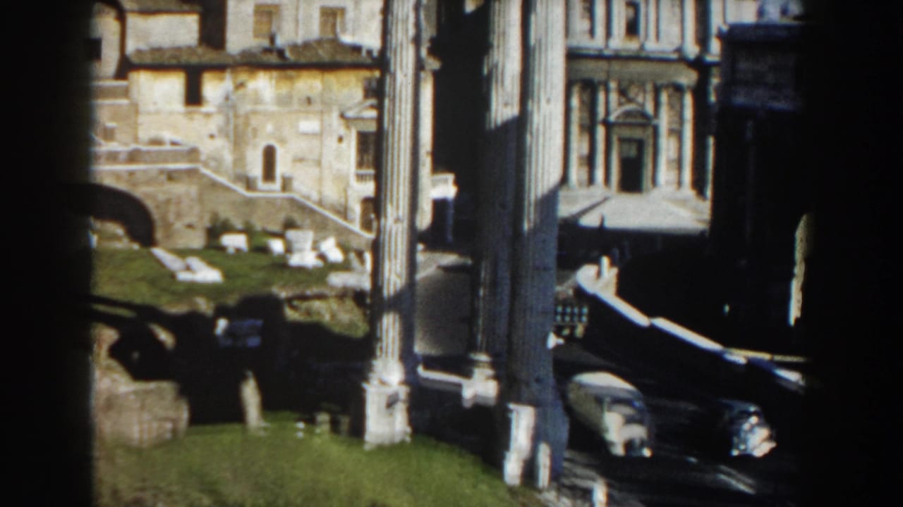 1959 Rome 8mm frame with tall columns in shadow, sunlit ruins, historic facades, grass, and light road traffic below.