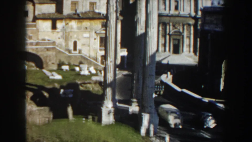 1959 Rome 8mm frame with tall columns in shadow, sunlit ruins, historic facades, grass, and light road traffic below.