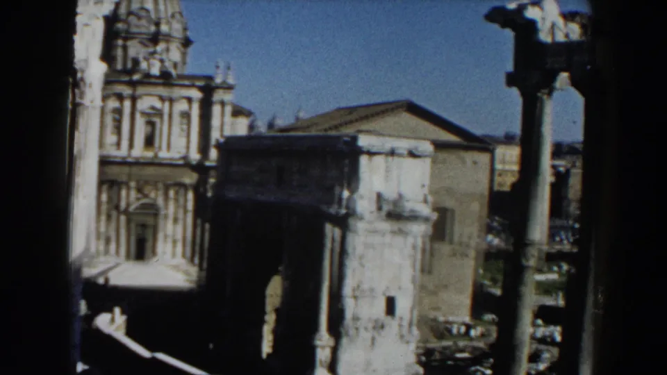 1959 Rome 8mm frame with a church-like facade, weathered tower, upright column, masonry ruins, and shadowed road area.