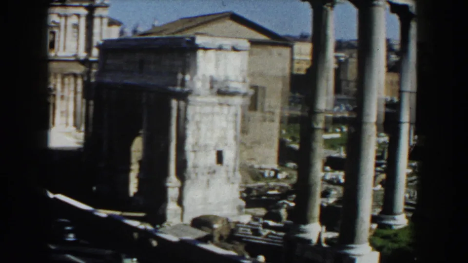 1959 8mm view in Rome showing ancient stone columns, ruin walls, grass, and surrounding city buildings under a blue sky.