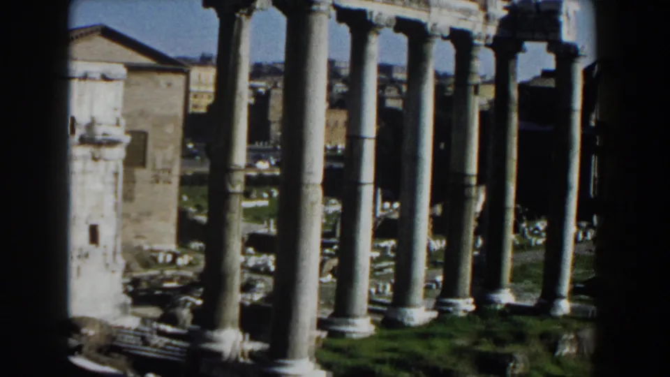 Rome 1959 home-movie frame with tall classical columns, pale stone ruins, grass, and distant urban structures in bright daylight.