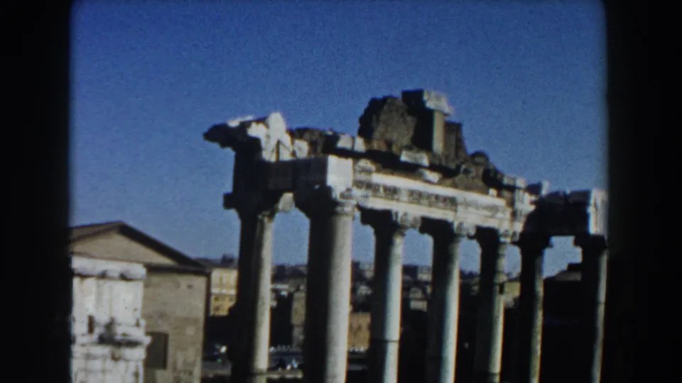 Rome 1959 8mm frame with classical columns, fragmented stonework, a nearby building, grass, and open blue sky.