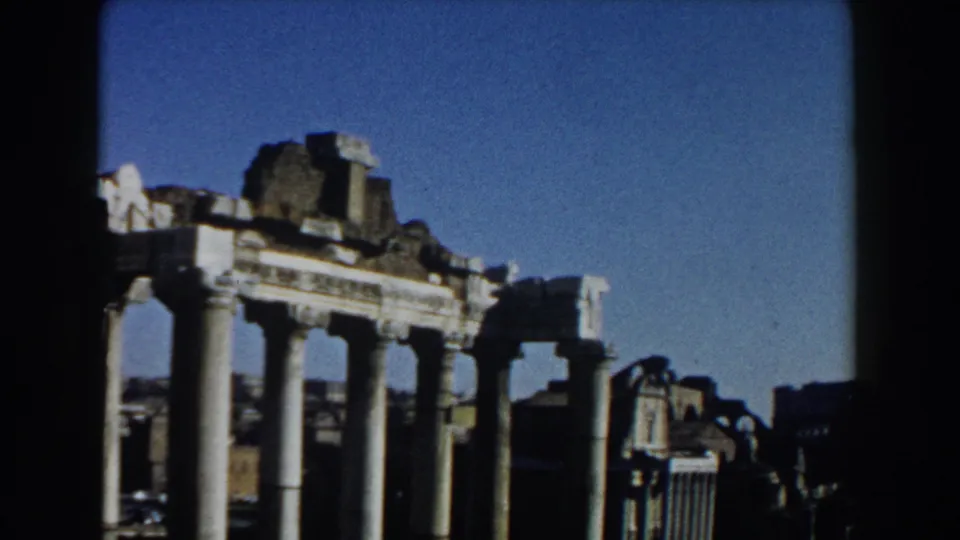 1959 Rome frame with ancient columns in the foreground and darker city buildings visible beyond the ruins.