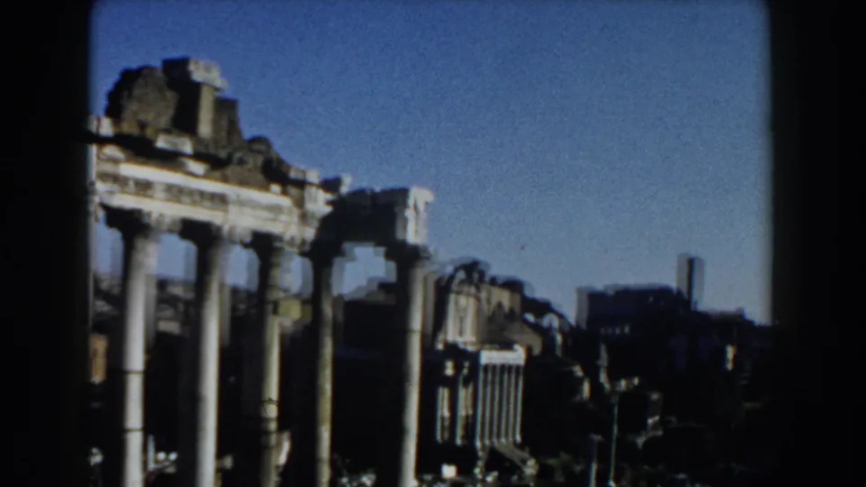 1959 Rome home-movie frame showing a panning view past pale columns, fragmented ruins, and a distant skyline.