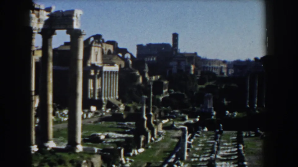 1959 Rome frame showing a wider view of ruins, standing columns, rubble-strewn ground, and city buildings in the background.