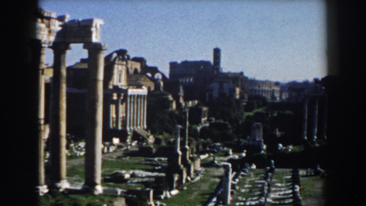 1959 8mm view across Roman ruins in Rome, with columns, stone structures, and distant historic architecture under bright sky.