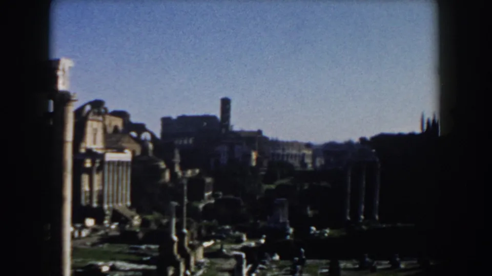 Rome home-movie frame showing ancient ruins, upright columns, and distant arched landmark architecture consistent with the Colosseum area.