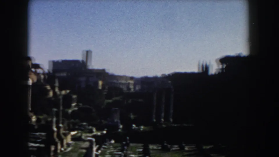 Soft 8mm frame from Rome showing columns, archaeological remains, distant buildings, and heavy shadow across the lower foreground.