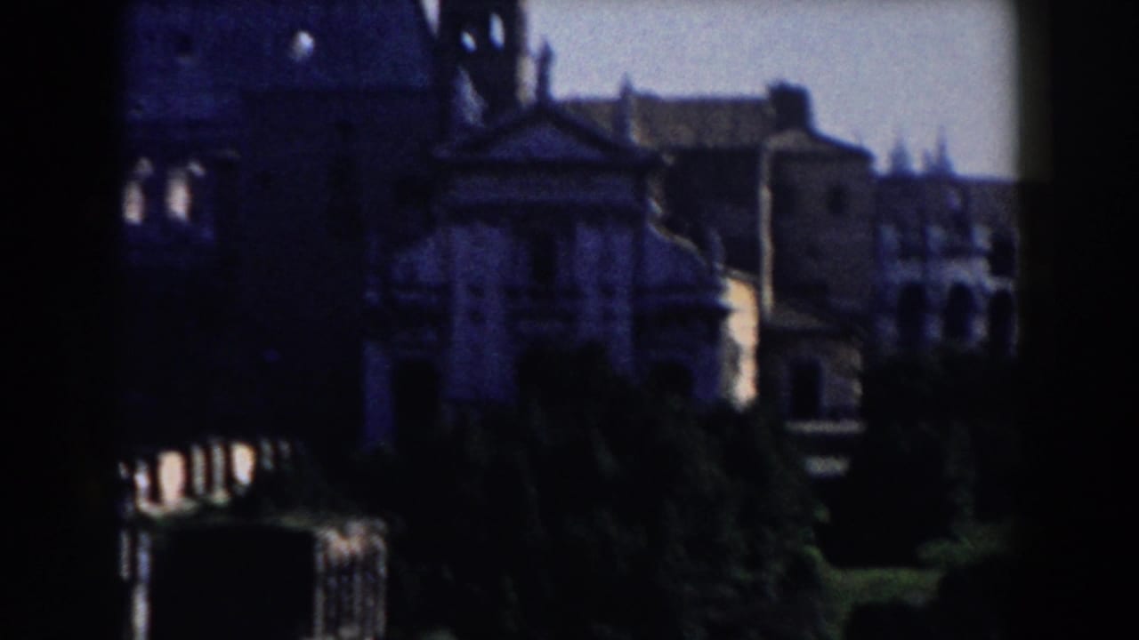 1959 Rome home-movie frame showing dark historic facades, a bell tower, trees, and shadowed foreground architecture.