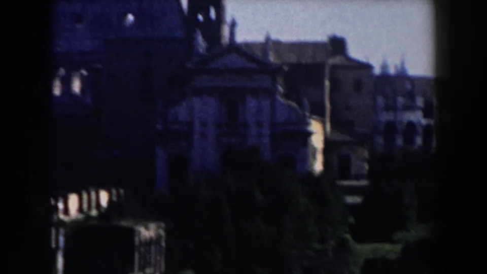 1959 Rome home-movie frame showing dark historic facades, a bell tower, trees, and shadowed foreground architecture.