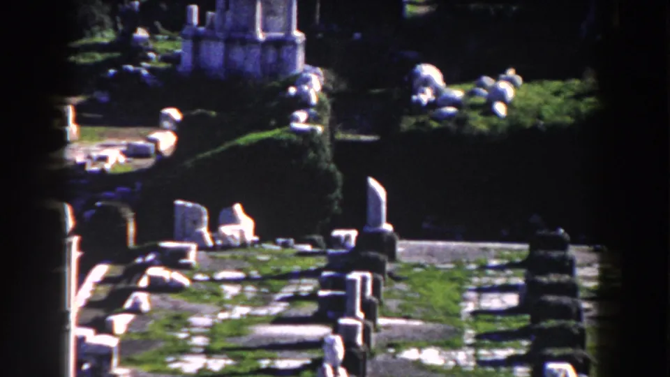 1959 Rome frame looking across a sunlit archaeological area with scattered stone slabs, upright markers, greenery, and low masonry.