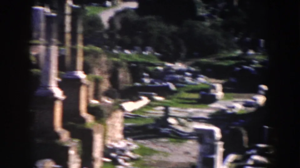 1959 Rome home-movie frame with vertical columns, low masonry, moss-covered foundations, and scattered fragments in soft focus.