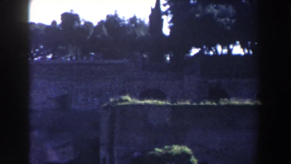 1959 Rome frame showing a darker section of the ruins with trees, a low stone wall, grass, and deep blue shadow from the film exposure.