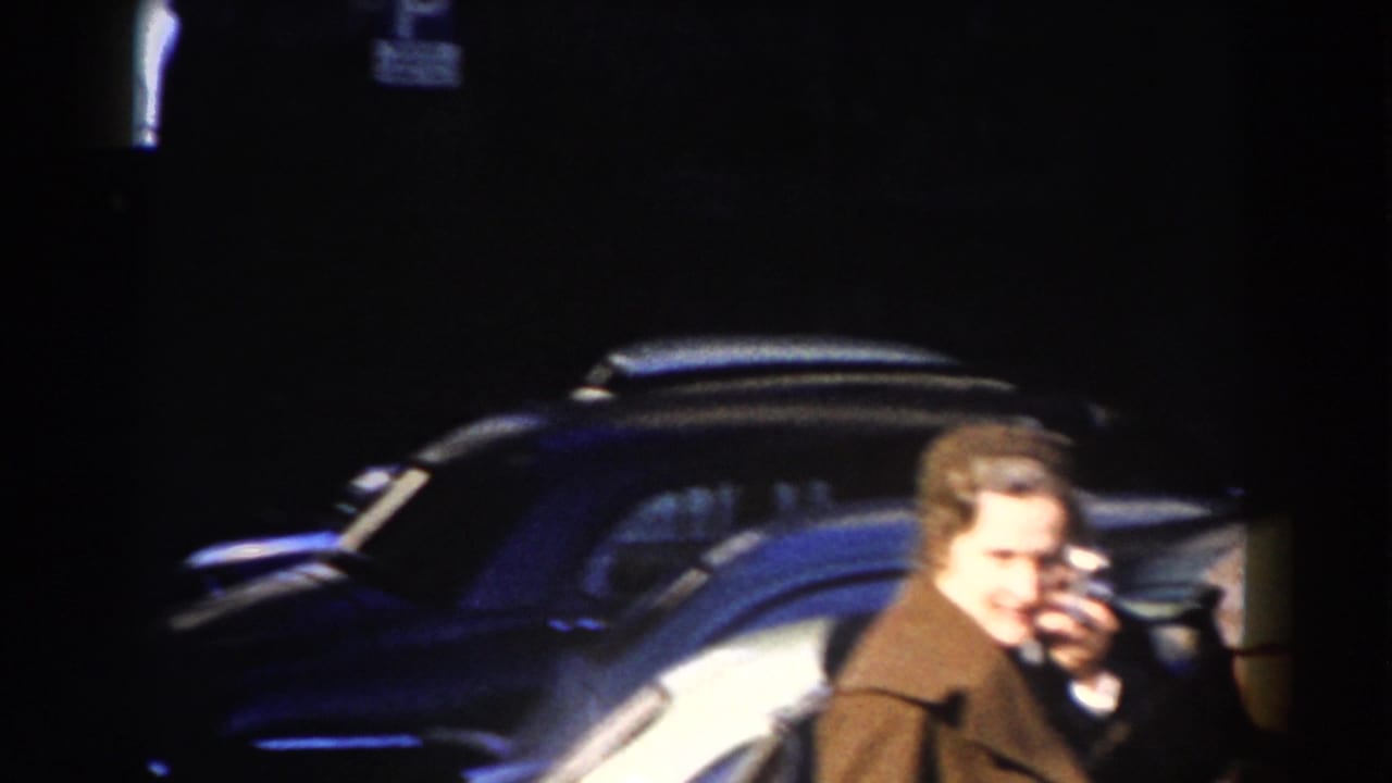 In Rome, a man in a brown overcoat stands near a parked mid-century car on a dark street, filmed in soft 8mm color.